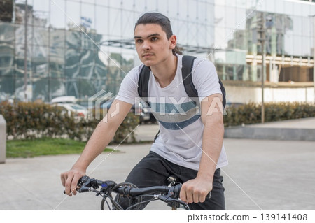 Young man riding a bicycle in an urban area near a modern building during daylight hours Young man riding a bicycle in an urban area near a modern building during daylight hours 139141408
