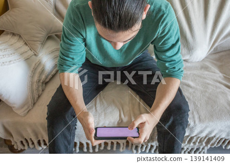 Young man sits on couch while using smartphone in a cozy indoor setting during daylight hours Young man sits on couch while using smartphone in a cozy indoor setting during daylight hours 139141409