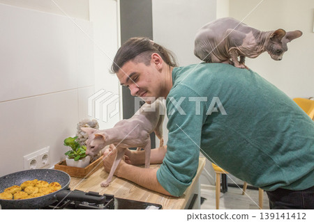 A man stands in a kitchen near a stove while a cat sits on his shoulder.  139141412