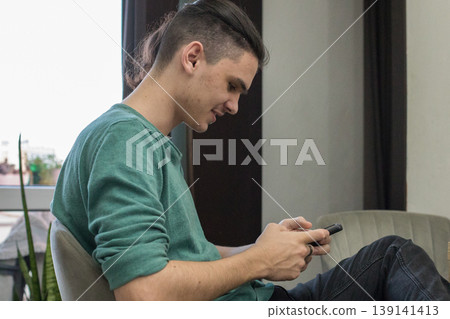 A young man is sitting in a chair and looking at his phone in a well-lit indoor area. A young man is sitting in a chair and looking at his phone in a well-lit indoor area. 139141413
