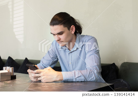 A man is seated at a table in a cafe. He holds a cup and appears to be lost in thought. 139141414