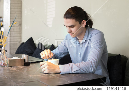 A man is seated at a table in a cafe. He holds a cup and appears to be lost in thought. 139141416