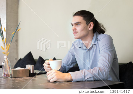 A young man is sitting at a table in a cafe. He looks at his phone and holds a cup i 139141417