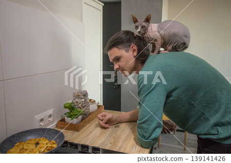 A man stands in a kitchen near a stove while a cat sits on his shoulder. A man stands in a kitchen near a stove while a cat sits on his shoulder. 139141426
