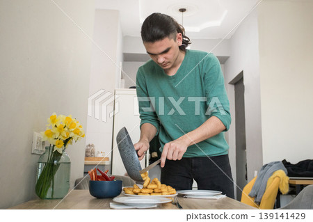 A young man serves food on a table in a kitchen. He holds a pan A young man serves food on a table in a kitchen. He holds a pan 139141429