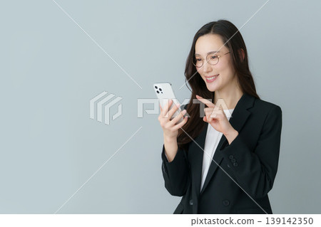 一位年輕的日本女性,身穿西裝,戴著灰色鏡框的眼鏡,手持智慧型手機。 一位年輕的日本女性,身穿西裝,戴著灰色鏡框的眼鏡,手持智慧型手機。 139142350