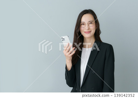 一位年輕的日本女性,身穿西裝,戴著灰色鏡框的眼鏡,手持智慧型手機。 一位年輕的日本女性,身穿西裝,戴著灰色鏡框的眼鏡,手持智慧型手機。 139142352