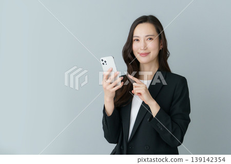 一位身穿西裝的年輕日本女性手持智慧型手機,背景為灰色。 一位身穿西裝的年輕日本女性手持智慧型手機,背景為灰色。 139142354