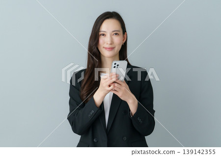 一位身穿西裝的年輕日本女性手持智慧型手機,背景為灰色。 一位身穿西裝的年輕日本女性手持智慧型手機,背景為灰色。 139142355