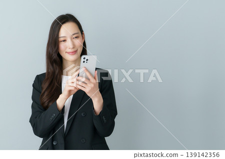 一位身穿西裝的年輕日本女性手持智慧型手機,背景為灰色。 一位身穿西裝的年輕日本女性手持智慧型手機,背景為灰色。 139142356