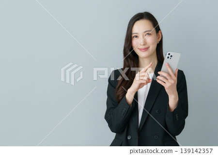 一位身穿西裝的年輕日本女性手持智慧型手機,背景為灰色。 一位身穿西裝的年輕日本女性手持智慧型手機,背景為灰色。 139142357