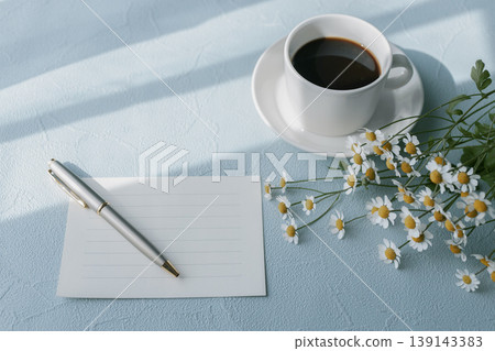 A morning scene image featuring coffee, matricaria, and a message card, on a blue background flat lay print. A morning scene image featuring coffee, matricaria, and a message card, on a blue background flat lay print. 139143383