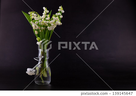 A bouquet of lilies of the valley on a black background with place for text. Delicate white spring flowers. Bouquet in a glass bottle. Copyspace. A bouquet of lilies of the valley on a black background with place for text. Delicate white spring flowers. Bouquet in a glass bottle. Copyspace. 139145647