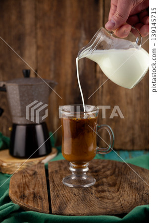 Hand Pouring Fresh Milk from Glass Jug into Coffee Cup with Moka Pot on Rustic Wooden Background Hand Pouring Fresh Milk from Glass Jug into Coffee Cup with Moka Pot on Rustic Wooden Background 139145715