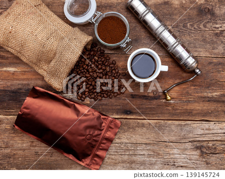 Bag of coffee on a wooden table. Coffee beans ground in a jar. Manual coffee grinder. Top view, place for text. Brown background. Packing coffee in a pack. White up with a drink. Bag of coffee on a wooden table. Coffee beans ground in a jar. Manual coffee grinder. Top view, place for text. Brown background. Packing coffee in a pack. White up with a drink. 139145724