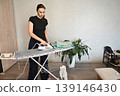 Woman ironing clothes at home in a cozy room with plants and warm lighting 139146430