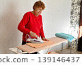 Middle age grey-haired woman talking on smartphone ironing clothes at laundry room 139146437