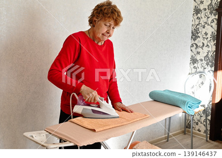 Middle age grey-haired woman talking on smartphone ironing clothes at laundry room Middle age grey-haired woman talking on smartphone ironing clothes at laundry room 139146437