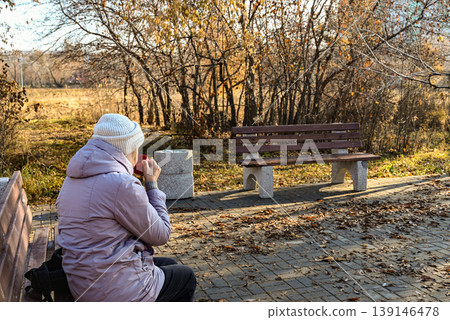 Older woman enjoys warm drink on a park bench during autumn afternoon 139146478