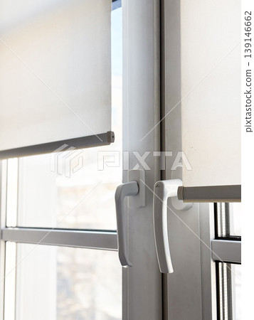 Two white window with handles and rolled blinds partially covering glass, closeup. Sunlight comes through, illuminating indoor space. Modern and clean setting. Daytime with clear visibility outside Two white window with handles and rolled blinds partially covering glass, closeup. Sunlight comes through, illuminating indoor space. Modern and clean setting. Daytime with clear visibility outside 139146662
