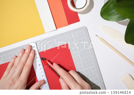 Hands using utility knife and ruler to cut red paper on cutting mat. Workspace for DIY paper crafts. 139146814