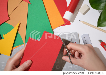 Hands cutting red paper with scissors on a creative workspace, colorful sheets and craft tools on desk. DIY, art, and handmade projects. Hands cutting red paper with scissors on a creative workspace, colorful sheets and craft tools on desk. DIY, art, and handmade projects. 139146816