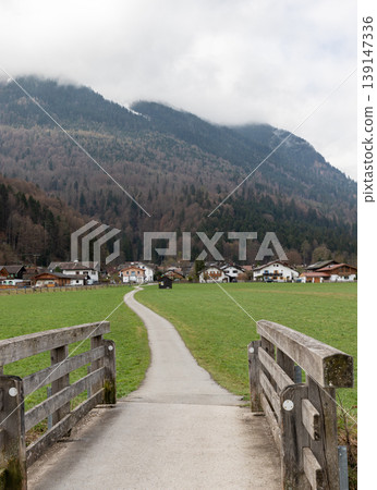 Rural path leading through green meadow toward alpine village with mountains and cloudy sky in Bavaria Germany Vertical Rural path leading through green meadow toward alpine village with mountains and cloudy sky in Bavaria Germany Vertical 139147336