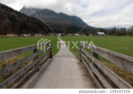 Rural path leading through green meadow toward alpine village with mountains and cloudy sky in Bavaria Germany Rural path leading through green meadow toward alpine village with mountains and cloudy sky in Bavaria Germany 139147339