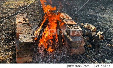 Small campfire with wooden sticks brightly burn at sunny summer day. Gentle flames and glowing embers create warm vibrant atmosphere. The scene conveys outdoor adventure and elemental beauty of fire Small campfire with wooden sticks brightly burn at sunny summer day. Gentle flames and glowing embers create warm vibrant atmosphere. The scene conveys outdoor adventure and elemental beauty of fire 139148153