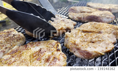 Male hands turning with plastic tongs pork steaks on home grill at sunny summer day. The focus is on the careful flipping and the rich texture of the meat as they cook evenly over the fire Male hands turning with plastic tongs pork steaks on home grill at sunny summer day. The focus is on the careful flipping and the rich texture of the meat as they cook evenly over the fire 139148162