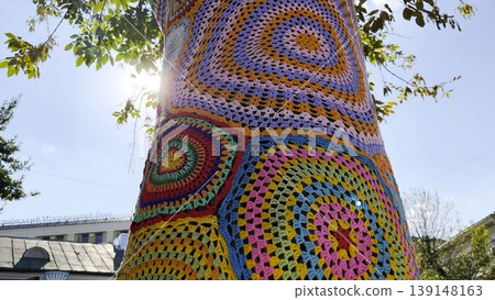 A tree trunk wrapped in colorful knitted cover stands on city street. Handmade yarn bombing installation transforms natural surface into vibrant piece of urban art, blending creativity and texture A tree trunk wrapped in colorful knitted cover stands on city street. Handmade yarn bombing installation transforms natural surface into vibrant piece of urban art, blending creativity and texture 139148163