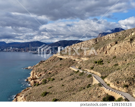 Spectacular coastal pathway showcasing rugged cliffs beside bustling city bay under dramatic clouds 139150328