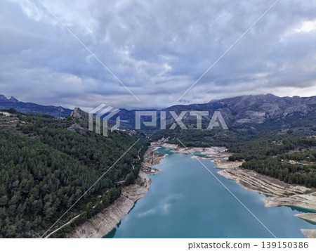Calm mountain water amid dense forest shadows, Serene lake scene with rocky edges and overcast skies 139150386