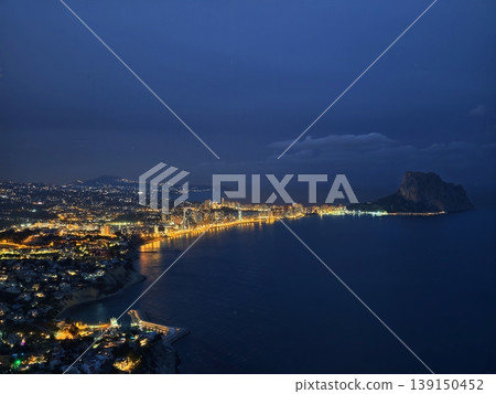Nighttime skyline featuring towering structures by glowing shoreline and tranquil dark sea 139150452
