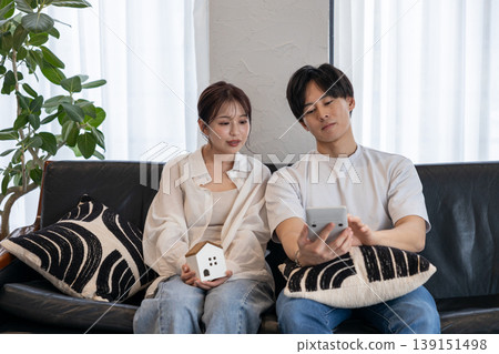 一對年輕夫婦坐在沙發上,手裡拿著工藝材料和計算器,討論他們的房屋建造計劃。 一對年輕夫婦坐在沙發上,手裡拿著工藝材料和計算器,討論他們的房屋建造計劃。 139151498