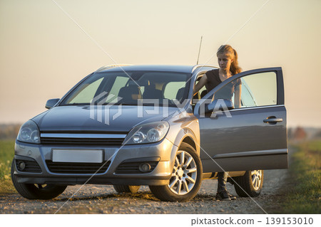 Young slim attractive long-haired woman in jeans and t-shirt at silver car with open door on empty gravel field road on clear bright sky copy space background. Problems and breakdowns concept. Young slim attractive long-haired woman in jeans and t-shirt at silver car with open door on empty gravel field road on clear bright sky copy space background. Problems and breakdowns concept. 139153010