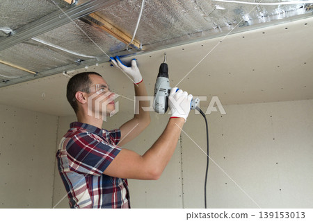 Young man in goggles fixing drywall suspended ceiling to metal frame using electrical screwdriver on ceiling insulated with shiny aluminum foil. Renovation, construction, do it yourself concept. Young man in goggles fixing drywall suspended ceiling to metal frame using electrical screwdriver on ceiling insulated with shiny aluminum foil. Renovation, construction, do it yourself concept. 139153013
