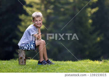 Young cute blond child boy sitting on tree stump on green grassy clearing on bright summer day. Young cute blond child boy sitting on tree stump on green grassy clearing on bright summer day. 139153015