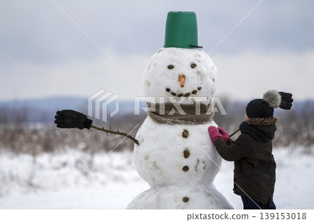 Young child boy finish doing smiling snowman in bucket hat, scarf and gloves on empty winter field landscape and clear blue sky copy space background. Merry Christmas, Happy New Year greeting card. Young child boy finish doing smiling snowman in bucket hat, scarf and gloves on empty winter field landscape and clear blue sky copy space background. Merry Christmas, Happy New Year greeting card. 139153018