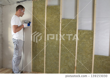 Worker in goggles with screwdriver working on insulation. Drywall on wall beams, insulating rock wool staff in wooden frame. Comfortable warm home, economy, construction and renovation concept. Worker in goggles with screwdriver working on insulation. Drywall on wall beams, insulating rock wool staff in wooden frame. Comfortable warm home, economy, construction and renovation concept. 139153030