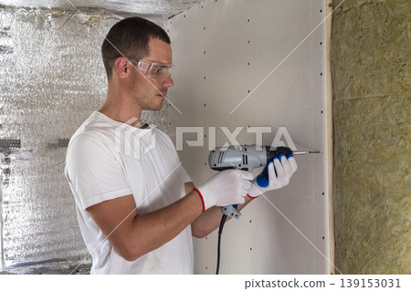 Worker in goggles with screwdriver working on insulation. Drywall on wall beams, insulating rock wool staff in wooden frame. Comfortable warm home, economy, construction and renovation concept. Worker in goggles with screwdriver working on insulation. Drywall on wall beams, insulating rock wool staff in wooden frame. Comfortable warm home, economy, construction and renovation concept. 139153031