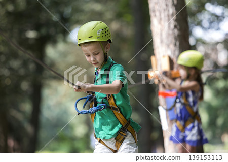 Two children, boy and girl in protective harness and safety helmets at climbing activity on rope way. Two children, boy and girl in protective harness and safety helmets at climbing activity on rope way. 139153113