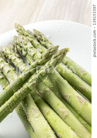 A photograph of spring vegetables, with fresh green asparagus arranged and glistening with water droplets. 139153397