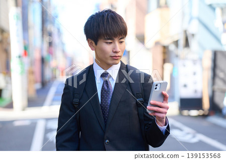 一位年輕的日本商人正在進行外出銷售,他一手拿著智慧型手機,走在城市街道上。 一位年輕的日本商人正在進行外出銷售,他一手拿著智慧型手機,走在城市街道上。 139153568