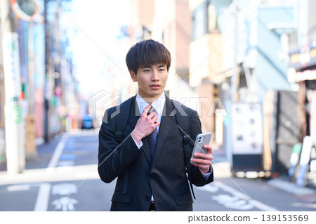 一位年輕的日本商人正在進行外出銷售,他一手拿著智慧型手機,走在城市街道上。 一位年輕的日本商人正在進行外出銷售,他一手拿著智慧型手機,走在城市街道上。 139153569