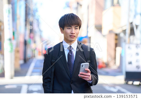 一位年輕的日本商人正在進行外出銷售,他一手拿著智慧型手機,走在城市街道上。 一位年輕的日本商人正在進行外出銷售,他一手拿著智慧型手機,走在城市街道上。 139153571