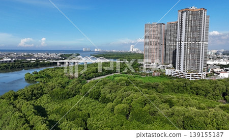 An aerial perspective captures a lush, dense mangrove forest split by a winding river, leading toward a distant urban skyline featuring white arched bridges under a vast, bright blue sky in Indonesia. 139155187