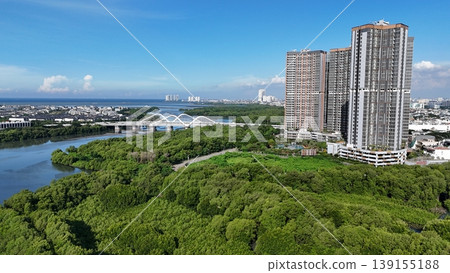 An aerial perspective captures a lush, dense mangrove forest split by a winding river, leading toward a distant urban skyline featuring white arched bridges under a vast, bright blue sky in Indonesia. An aerial perspective captures a lush, dense mangrove forest split by a winding river, leading toward a distant urban skyline featuring white arched bridges under a vast, bright blue sky in Indonesia. 139155188