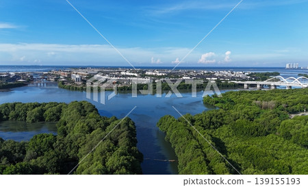 An aerial perspective captures a lush, dense mangrove forest split by a winding river, leading toward a distant urban skyline featuring white arched bridges under a vast, bright blue sky in Indonesia. 139155193