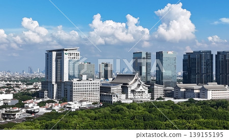 An aerial perspective captures a lush, dense mangrove forest split by a winding river, leading toward a distant urban skyline featuring white arched bridges under a vast, bright blue sky in Indonesia. 139155195
